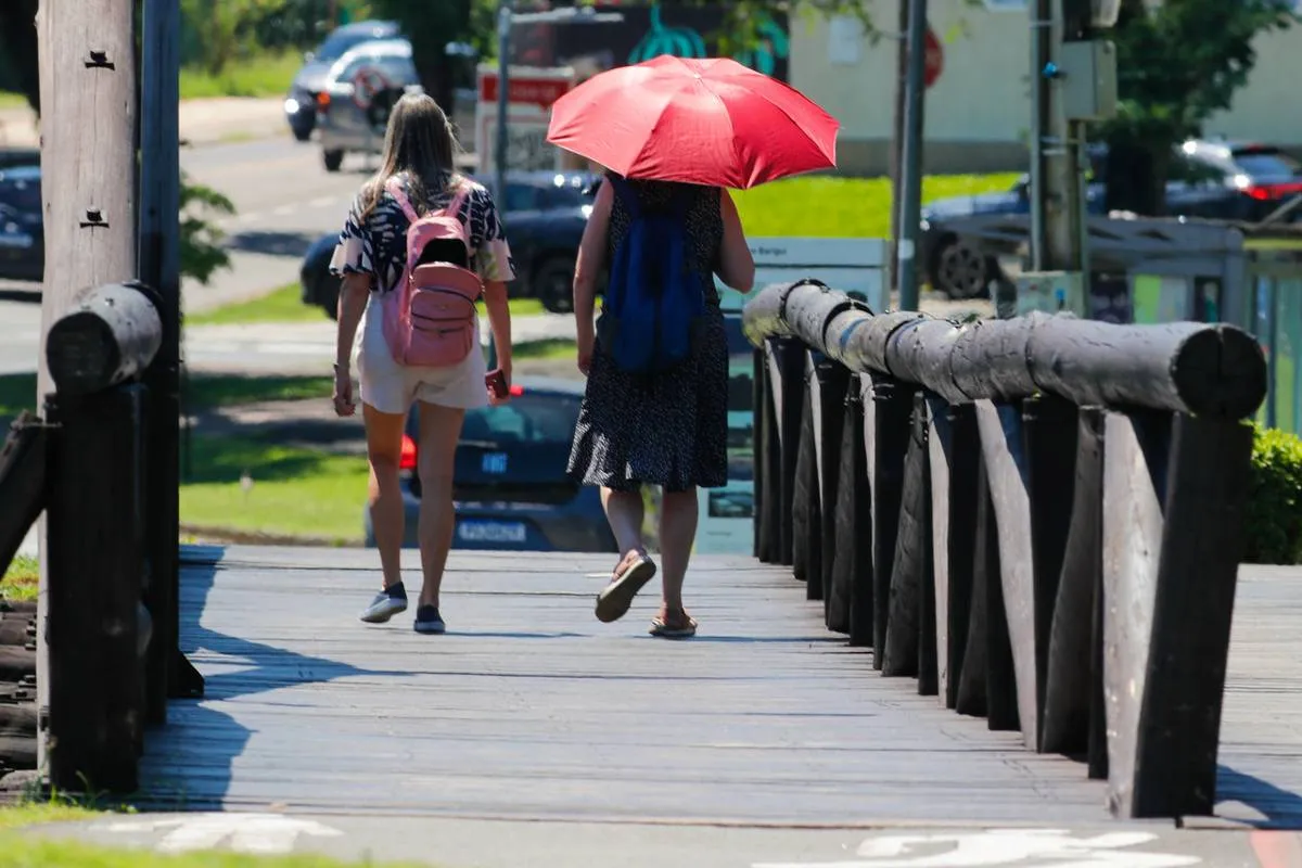 Imagem mostra pessoas com calor andando em um parque. Uma pessoa se protege do sol com um guarda chuva.