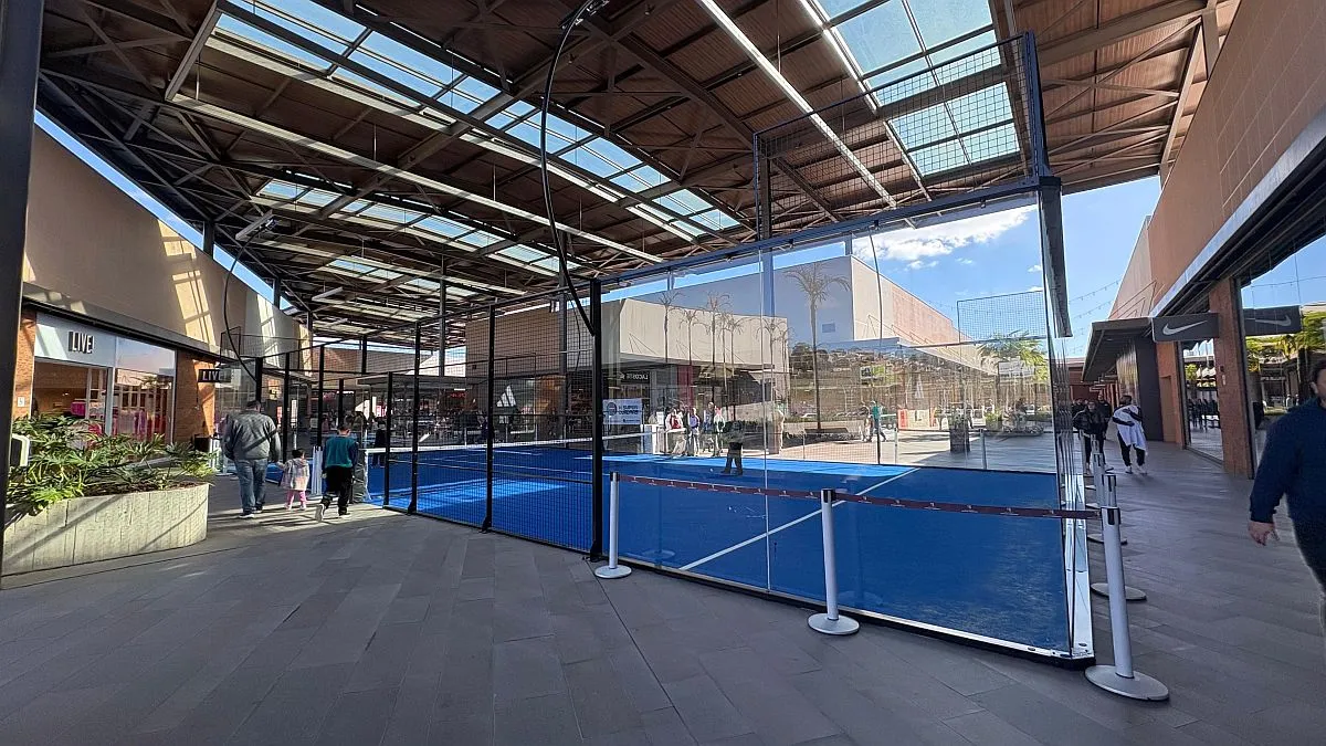 imagem mostra área central de Outlet com uma quadra de padel montada. Pessoas passam ao redor e lojas compõem o cenário