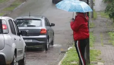 Imagem mostra uma mulher com calça preta, blusa vermelha e um guarda chuva azul na beira de uma rua com poça de água.