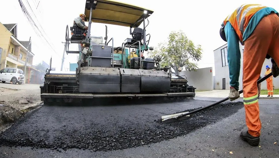 Imagem mostra trabalhador puxando asfalto que uma grande máquina despeja na rua