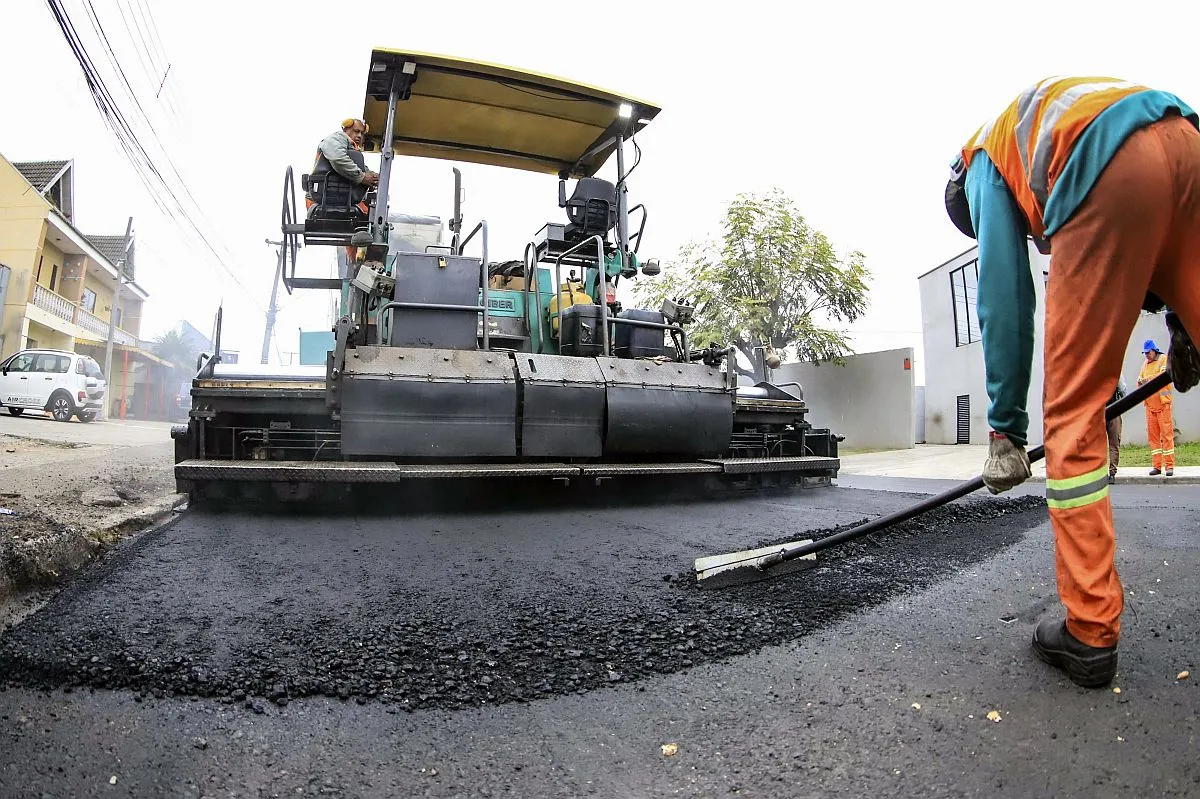 Imagem mostra trabalhador puxando asfalto que uma grande máquina despeja na rua