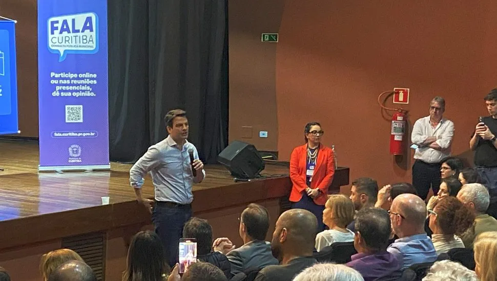 imagem mostra prefeito de Curitiba discrusando para várias pessoas em um auditório, Um banner com a frase Fala Curitiba é destaque