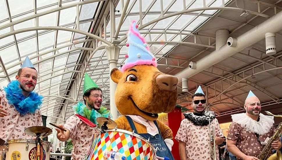 Imagem mostra mascote capivara no hall do shopping, com músicos vestidos de pierrôt com instrumentos musicais