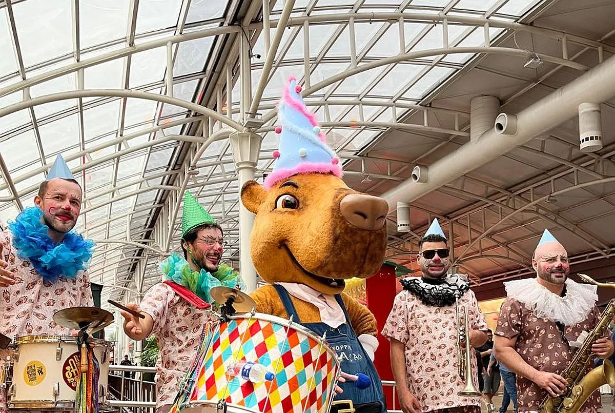 Imagem mostra mascote capivara no hall do shopping, com músicos vestidos de pierrôt com instrumentos musicais