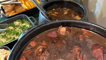 Em primeiro plano uma panelada de feijoada, com outros pratos ao fundo, desfocados