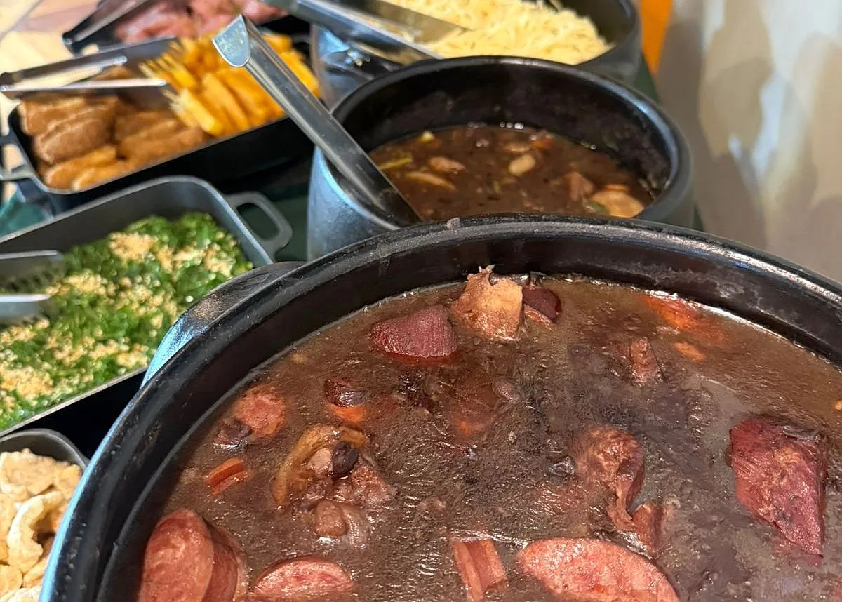 Em primeiro plano uma panelada de feijoada, com outros pratos ao fundo, desfocados