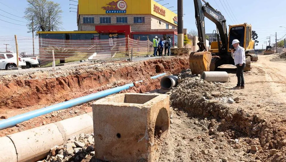 imagem mostra rua aberta com tubulação de água exposta. Há uma escavadeira em primeiro plano e alguns dos funcionários da empresa qe realiza o serviço
