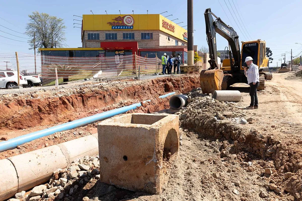 imagem mostra rua aberta com tubulação de água exposta. Há uma escavadeira em primeiro plano e alguns dos funcionários da empresa qe realiza o serviço