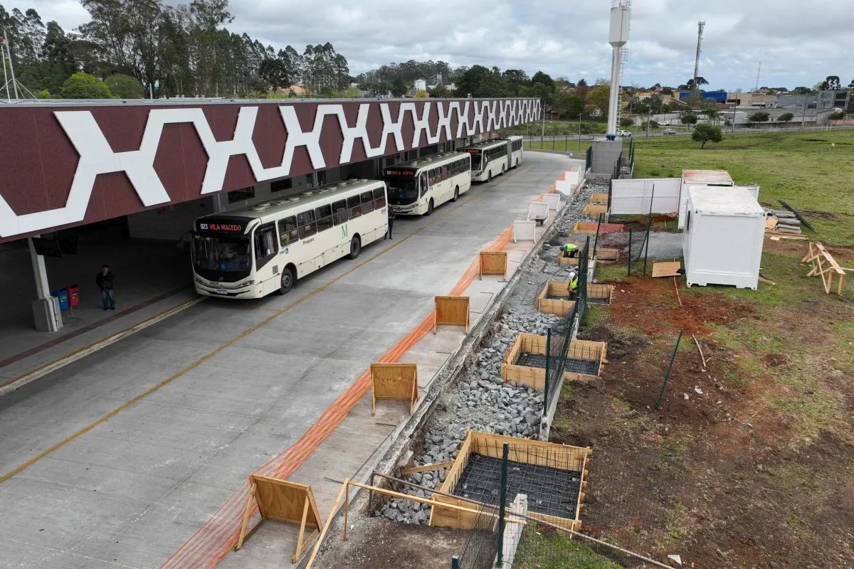 Imagem aérea mostra no lado esquerdo três ônibus enfileirados e do lado direito da rua de acesso o canteiro de obras