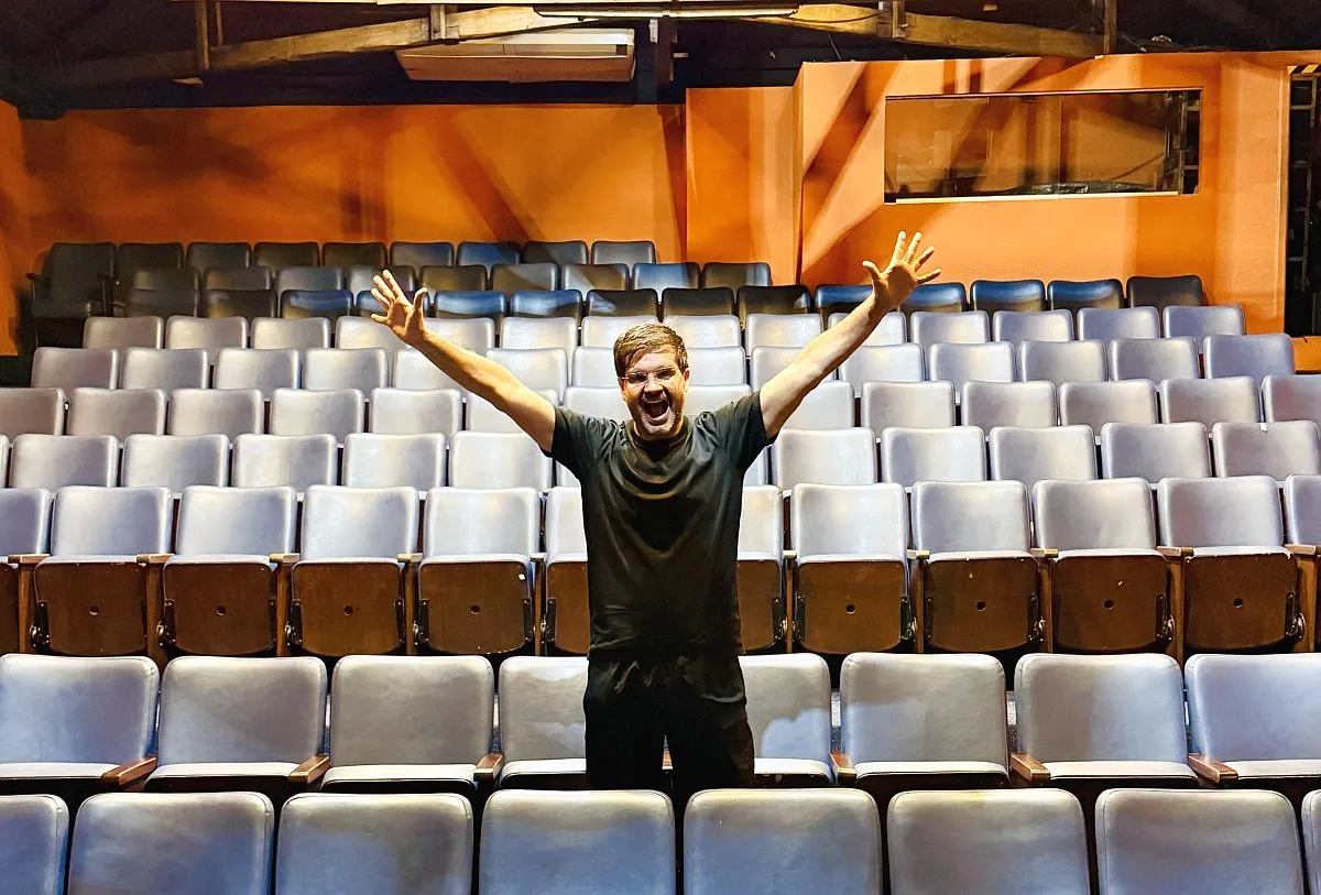 Imagem mostra Ilan, o Moço do Te Amo, no meio do auditório do teatro Barracão Encena.