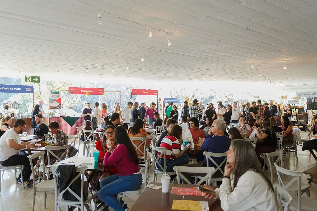 Imagem mostra várias mesas com convidados no hall de entrada do MON. Ao fundo, barraquinhas de expositores
