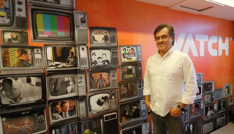 Imagem mostr ao personagem entrevistado pela matéria em frente a um painel com várias imagens de televisores antigos. Ele veste camisa branca e calça jeans