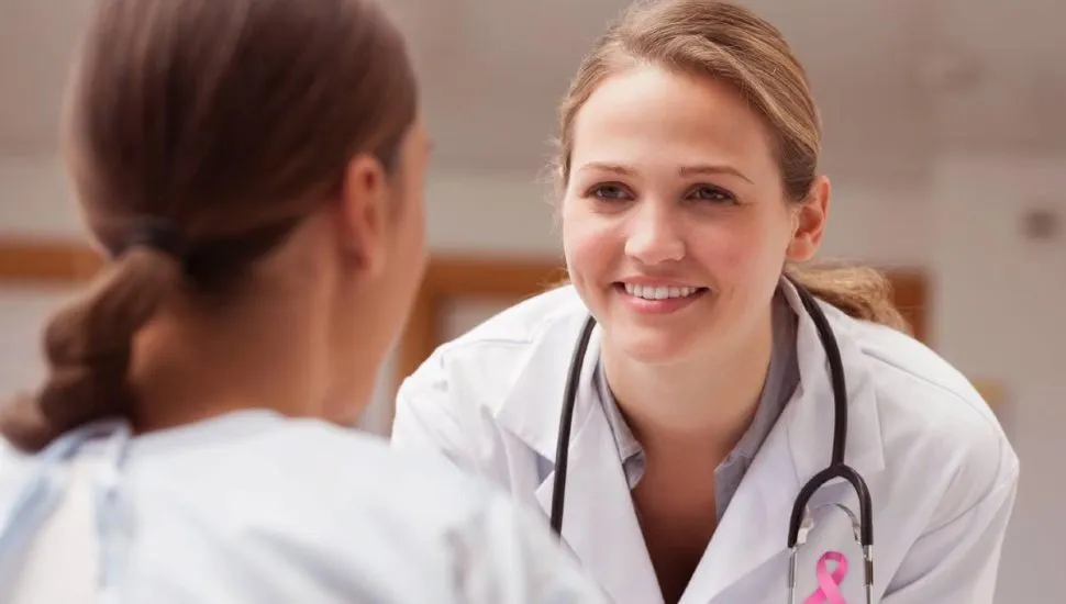Imagem mostra uma médica conversando com uma paciente. A médica tem no jaleco um símbolo da luta contra o câncer de mama.