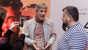 ator Roney Vilela dá entrevista em frente a um cartaz do filme que ele participa, com repórter ao seu lado e uma mulher filmando tudo com celular