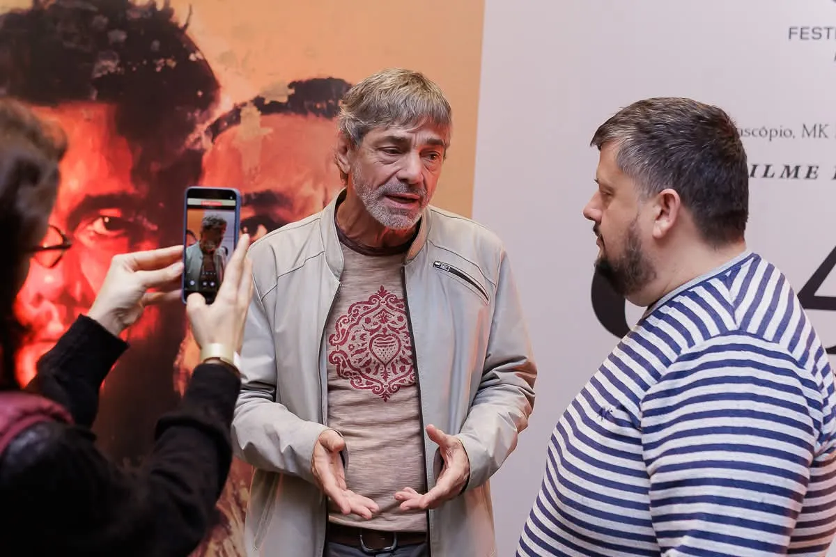 ator Roney Vilela dá entrevista em frente a um cartaz do filme que ele participa, com repórter ao seu lado e uma mulher filmando tudo com celular