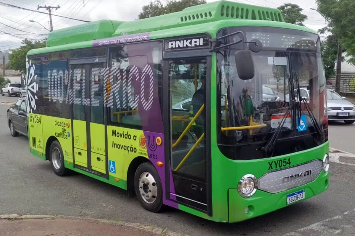 Imagem mostra um micro ônibus elétrico  de cor verde flagrado circulando pelas ruas de Curitiba em uma linha de transporte coletivo.