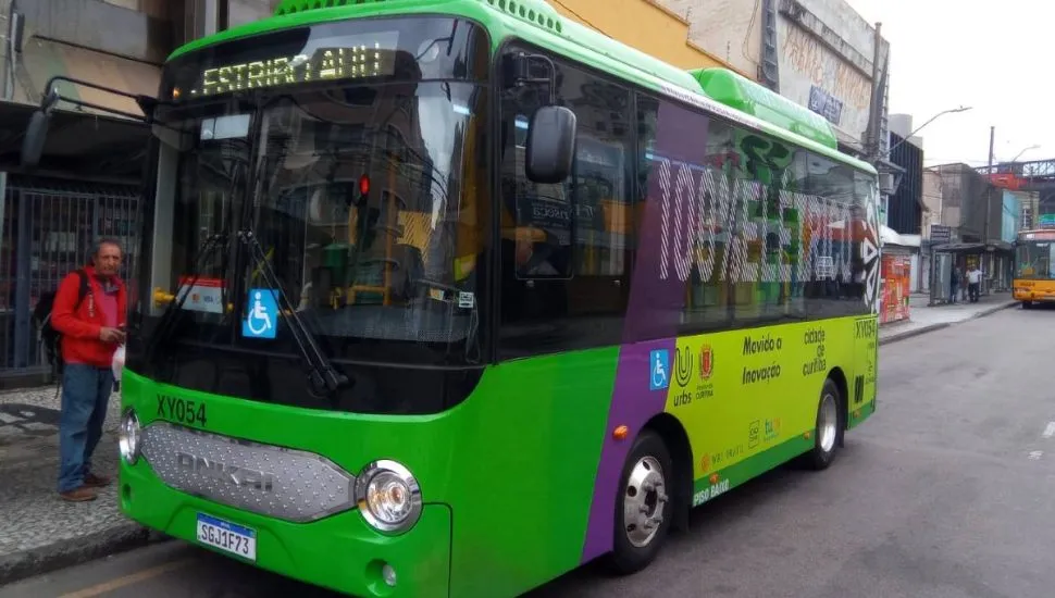 Imagem mostra um micro ônibus elétrico de cor verde flagrado circulando pelas ruas de Curitiba em uma linha de transporte coletivo.