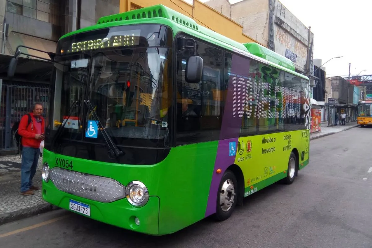Imagem mostra um micro ônibus elétrico  de cor verde flagrado circulando pelas ruas de Curitiba em uma linha de transporte coletivo.