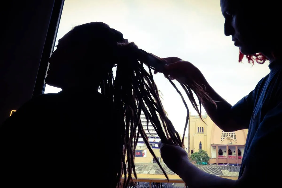 Imagem contra a luz mostra uma pessoa fazendo dreadlocks em outra.