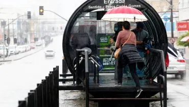 Imagem mostra uma mulher entrando em uma estação-tubo com muita chuva em Curitiba