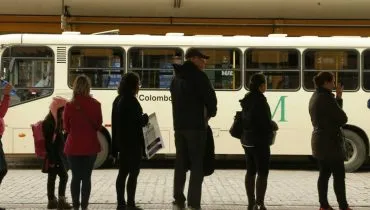 Imagem mostra passageiros em uma fila esperando o ônibus.