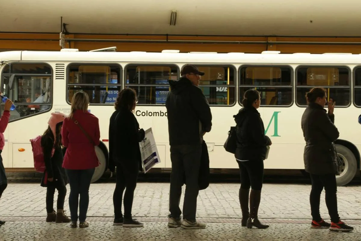 Imagem mostra passageiros em uma fila esperando o ônibus.