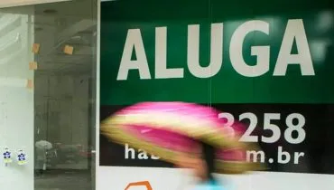 Imagem mostra uma pessoa passando na frente uma placa de aluga no Centro de Curitiba. está chovendo e a pessoa está de guarda-chuva