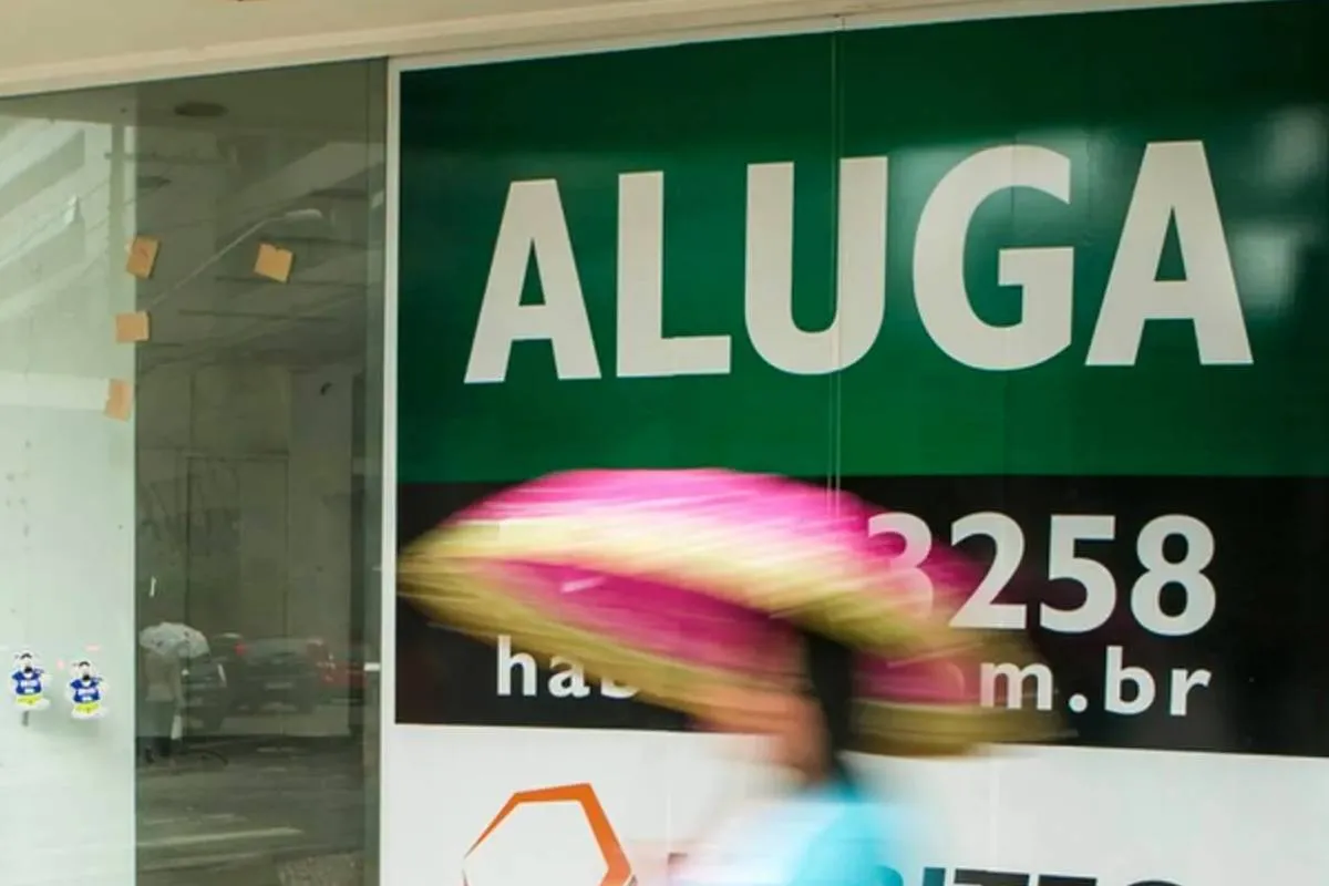 Imagem mostra uma pessoa passando na frente uma placa de aluga no Centro de Curitiba. está chovendo e a pessoa está de guarda-chuva