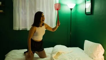Imagem mostra uma mulher em uma cama tentando espantar/matar um mosquito à noite.