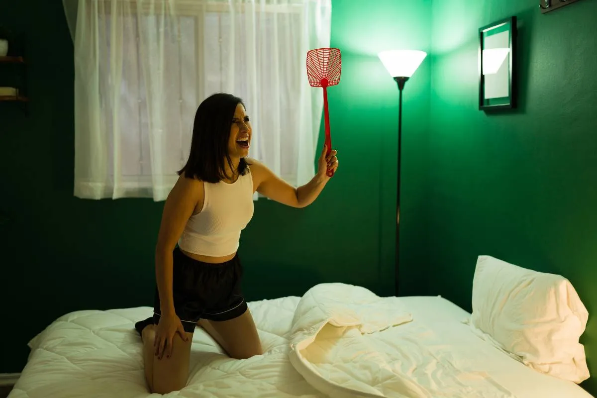 Imagem mostra uma mulher em uma cama tentando espantar/matar um mosquito à noite.