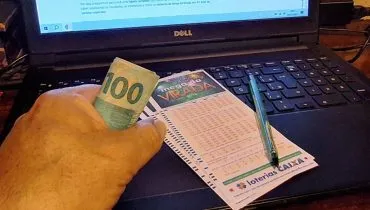 Imagem mostra uma pessoa com uma nota de 100 reais na mão ao lado de bilhete de apostas da Mega da Virada.
