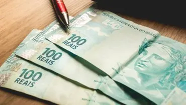 Imagem mostra três notas de cem reais em cima de uma mesa.