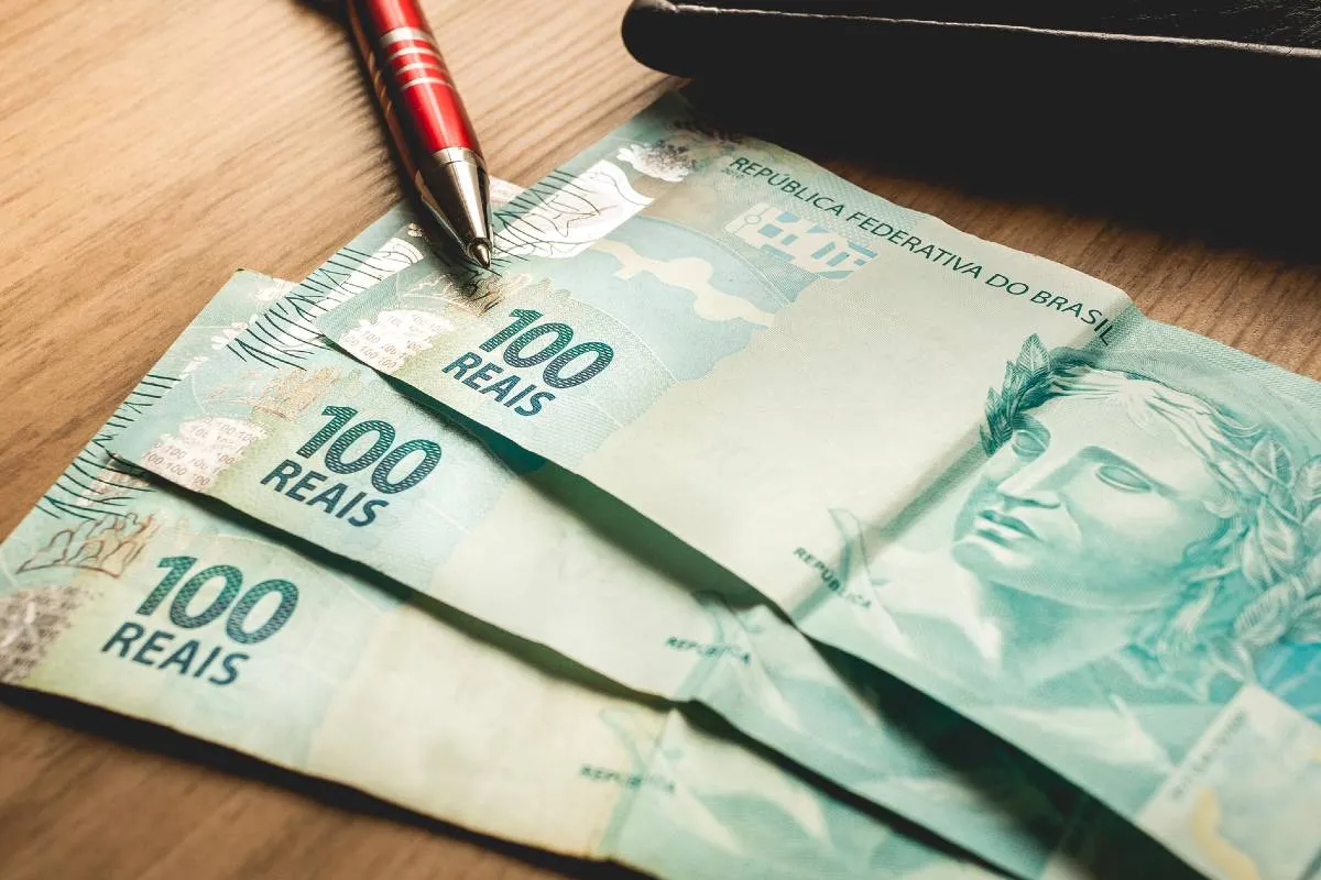Imagem mostra três notas de cem reais em cima de uma mesa.