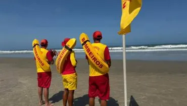 Imagem mostra três Bombeiros olhando para o mar ao lado de uma bandeira amarela.