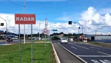 Imagem mostra uma rua com uma placa de radar ao centro.