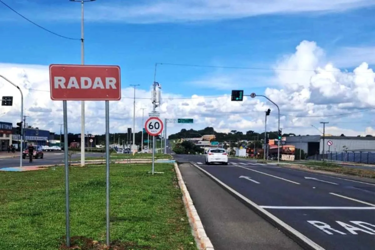 Imagem mostra uma rua com uma placa de radar ao centro.