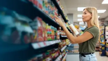 Imagem mostra uma mulher em uma prateleira escolhendo produtos.