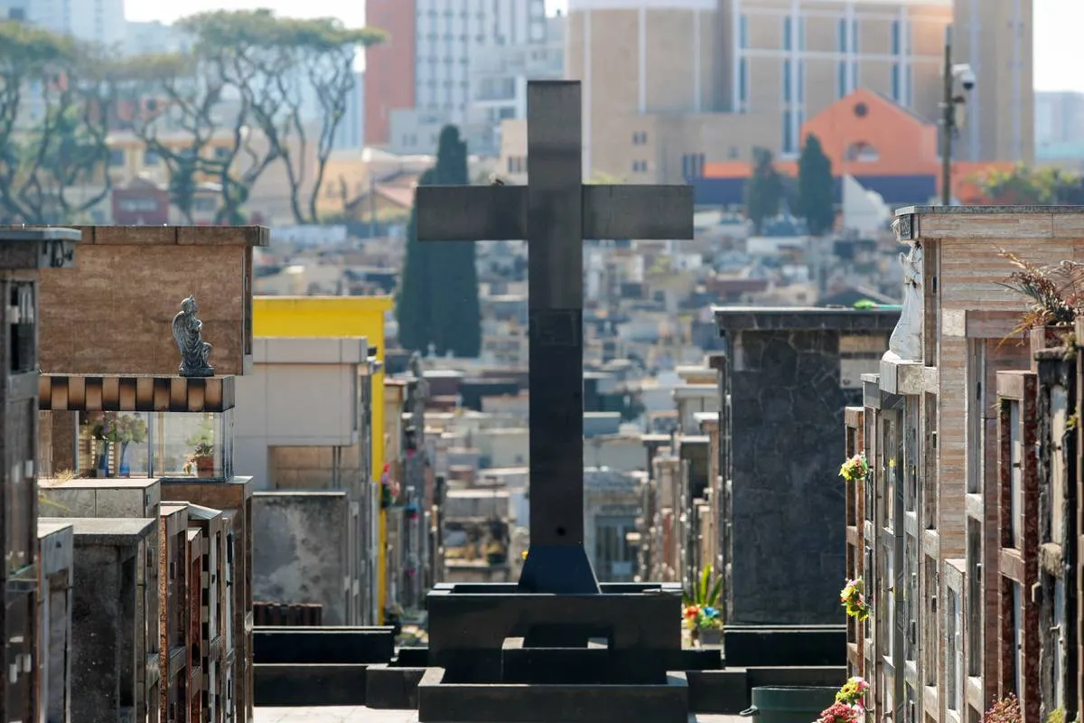 Imagem mostra um detalhe de um cemitério em Curitiba, com uma cruz e túmulos.