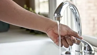 imagem mostra mão de homem fechando uma torneira que escorre água em uma cozinha