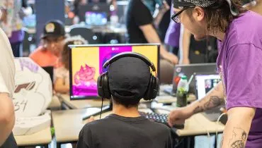 Imagem mostra um jovem com fones de ouvido, em frente a um computador, com um adulto ao seu lado passando orientações