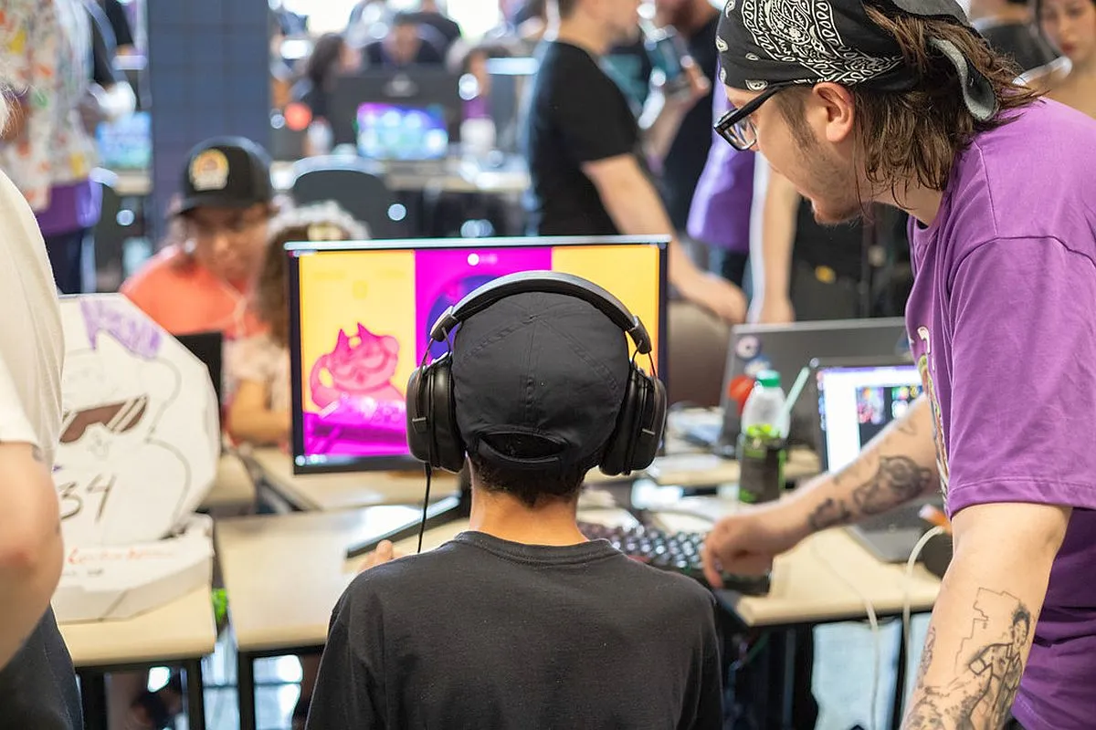 Imagem mostra um jovem com fones de ouvido, em frente a um computador, com um adulto ao seu lado passando orientações