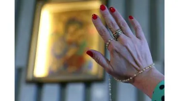 Imagem mostra a mão de uma mulher com o terço na mão e uma imagem religiosa ao fundo.