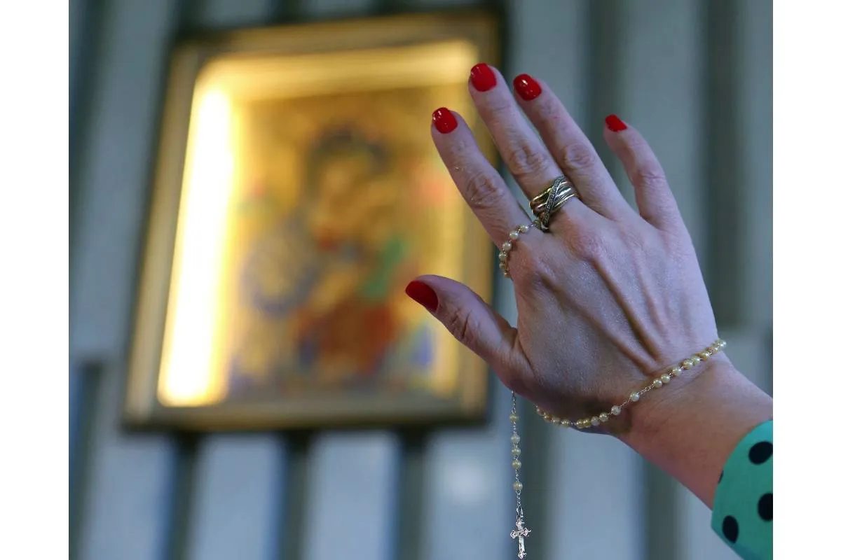 Imagem mostra a mão de uma mulher com o terço na mão e uma imagem religiosa ao fundo.