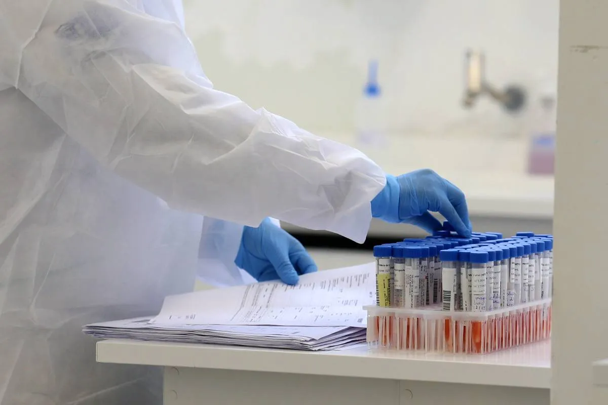 Imagem mostra exames de laboratório e uma pessoa protegida com uma roupa branca fazendo anotações.