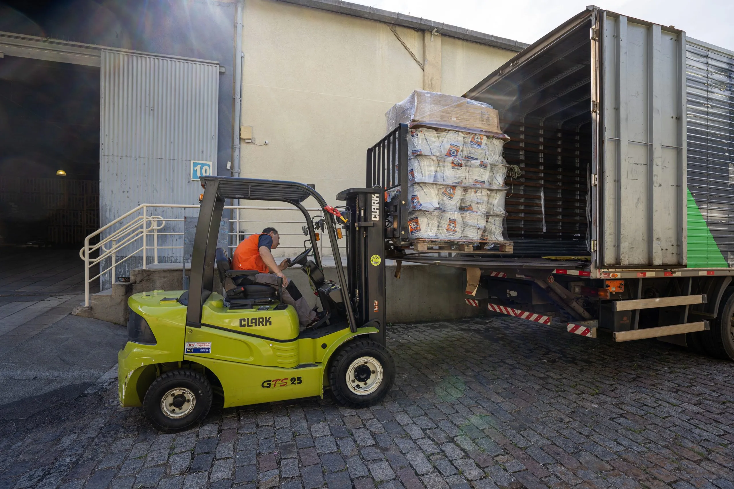 Imagem mostra uma empilhadeira verde carregando mantimentos em um caminhão.