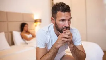 Imagem mostra um homem sentado em uma cama e uma mulher ao fundo.