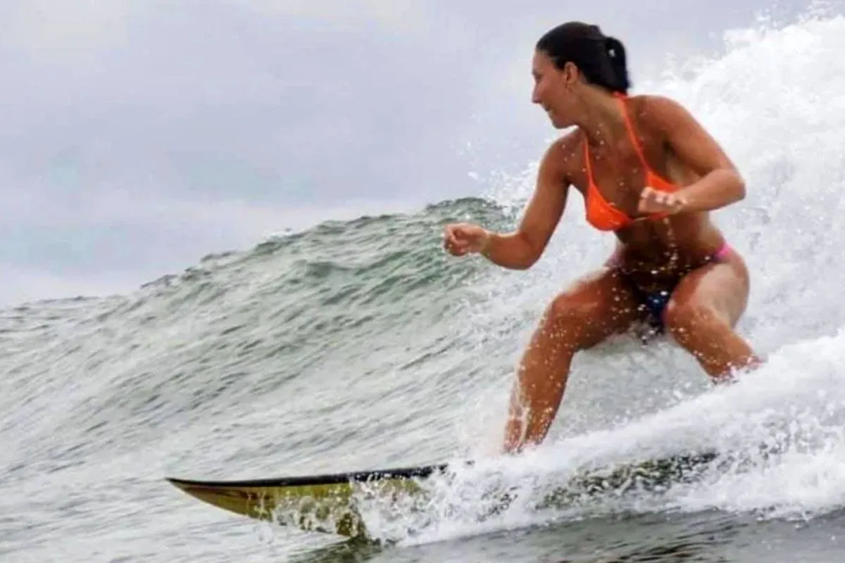 Imagem mostra uma mulher de cabelo escuro preso num rabo de cavalo, surfa de cócoras, numa onda. Ela está voltada para a direita, com o corpo ligeiramente inclinado para a frente e os braços abertos para o equilíbrio. Veste um biquíni cor-de-laranja vivo, com cuecas de biquíni azuis por baixo. A prancha de surf é amarela e verde. A onda é grande e verde-escura, com espuma branca a rebentar no cume e à volta da prancha de surf. A água está agitada e com espuma, com salpicos de água a voar no ar. O céu está encoberto e cinzento. A imagem é captada de um ângulo ligeiramente baixo, com a mulher e a prancha de surf a ocuparem a maior parte do enquadramento. A focagem é nítida na mulher e na prancha de surf, com a onda e o céu desfocados. A iluminação é natural e difusa, com cores vivas e realistas.
