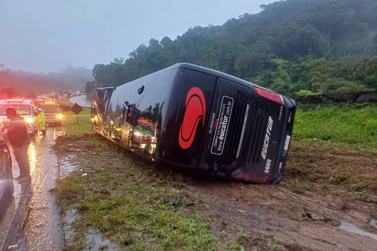 Imagem mostra um ônibus preto tombado ao lado da BR-116. Há neblina e carros com as luzes acesas em um congestionamento.