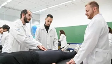 Imagem mostra fisioterapeutas em uma sessão com uma paciente. Eles vestem jalecos branco e a paciente está com roupa preta e cinza em uma maca.