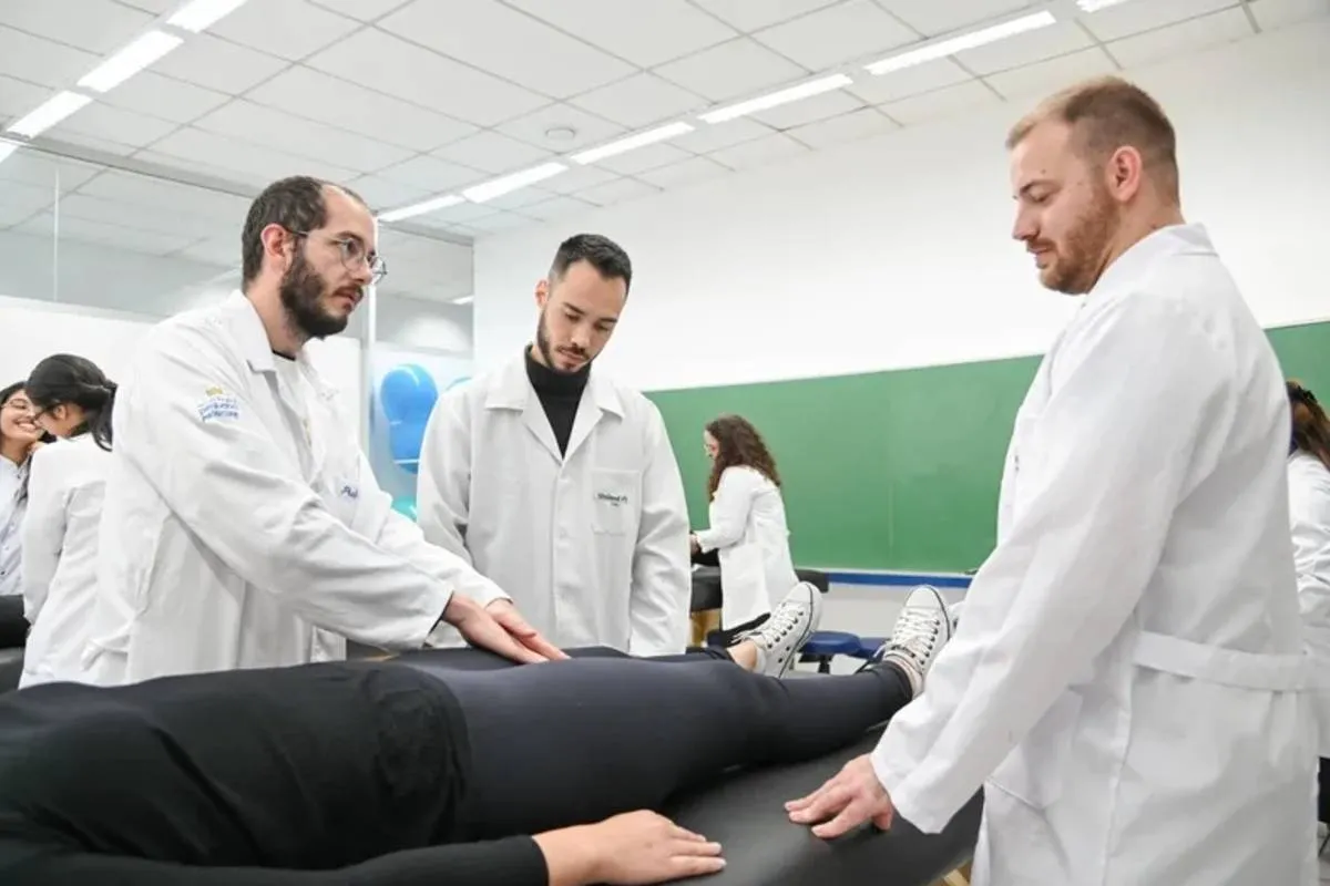 Imagem mostra fisioterapeutas em uma sessão com uma paciente. Eles vestem jalecos branco e a paciente está com roupa preta e cinza em uma maca.